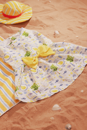A colorful beach dress with a yellow bow and matching striped hat lies on sandy beach alongside a striped towel and seashells.